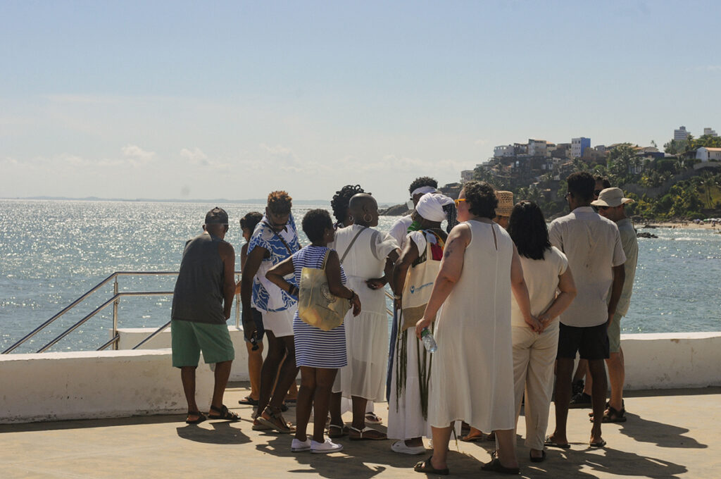 Grupo de pessoas reunido à beira-mar na Casa de Iemanjá , Rio Vermelho, durante um passeio guiado prévio a Festa de Iemanjá 2025. Clima de troca e escuta, em cenário ensolarado. Ao fundo o Morro da Paciência
